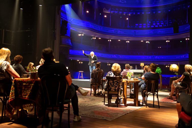 Een komiek staat op het podium van een theater, terwijl het publiek aan tafels toekijkt.