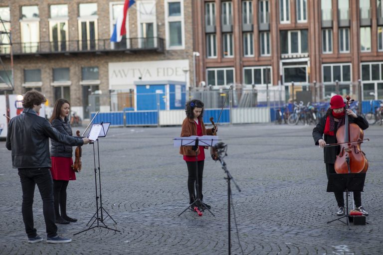 Drie muzikanten spelen op een plein, met een focus op hun instrumenten en bladmuziek.