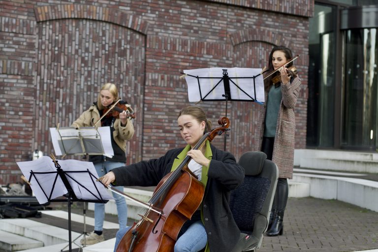 Drie muzikanten spelen samen op straat, met een cellist vooraan en twee violisten op de achtergrond.