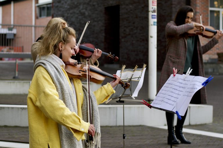 Een muzikant in een gele jas speelt viool buiten, terwijl een andere muzikant achter haar op een podium staat.
