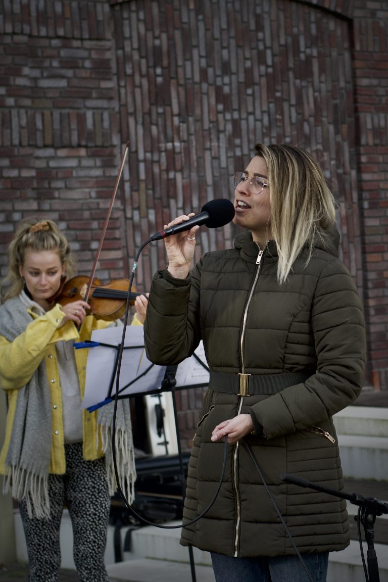 Een vrouw zingt in een microphone terwijl een meisje op de achtergrond een viool speelt.