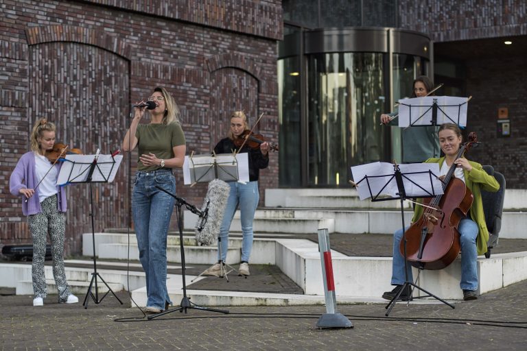 Een vrouwelijke zangeres met een microfoon staat op een podium, omringd door muzikanten met verschillende instrumenten.