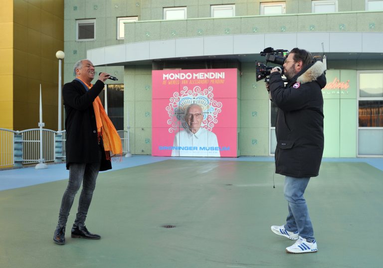 Een persoon zingt terwijl een cameraman hem filmt, met een kleurrijke achtergrond en een afbeelding van een man in het posterformaat.
