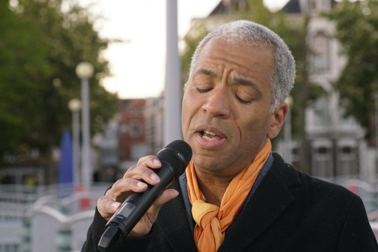 Een man zingt met een microfoon en heeft een oranje sjaal om zijn hals.
