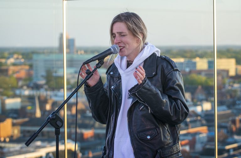 Een persoon met een leren jack en een hoodie staat op een podium en zingt in een microfoon met een stadslandschap op de achtergrond.