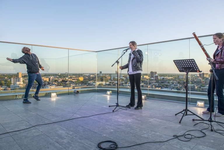 Een muzikant staat op een dakterras met een panoramisch uitzicht, terwijl hij in de microfoon zingt en wordt vergezeld door een andere muzikant.