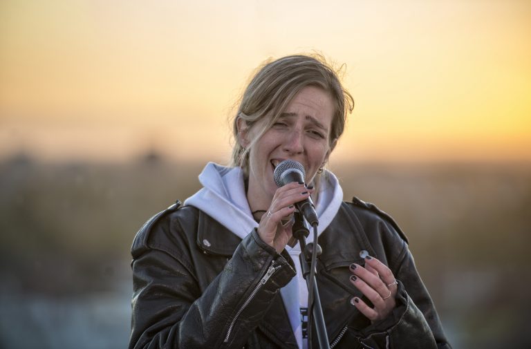 Een zangeres die een nummer vertolkt met een microfoon, tegen een achtergrond van een zonsondergang.