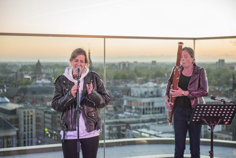 Twee muzikanten optreden op een hoog dak met een stadszicht op de achtergrond tijdens zonsondergang.