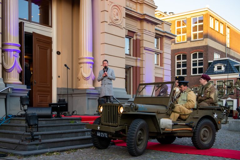 Een vintage jeep staat op een rode loper voor een gebouw, terwijl iemand op de trap een toespraak houdt.