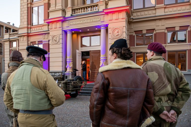 Drie mensen in historische kleding staan voor een groot gebouw met kleurrijke verlichting en een rode loper.