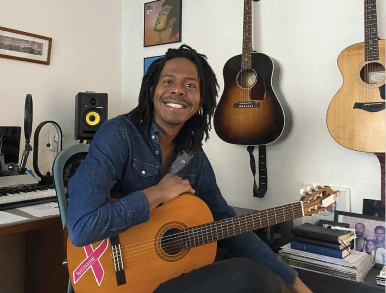 Een glimlachende man met een gitaar zit in een muziekstudio omringd door diverse muziekinstrumenten.