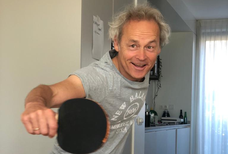 Een man met krullend haar lacht terwijl hij een tafeltennisbatje vasthoudt in een lichte binnenruimte.