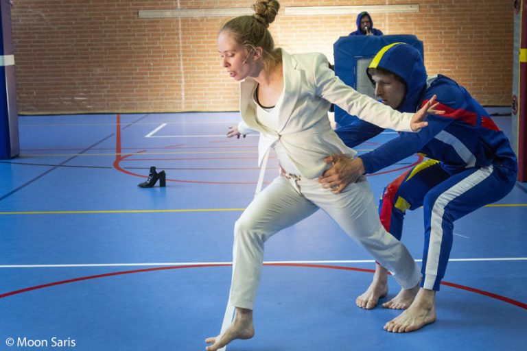 Een vrouw in een lichte outfit rent weg terwijl iemand in beschermende kleding haar probeert vast te houden in een sportzaal.