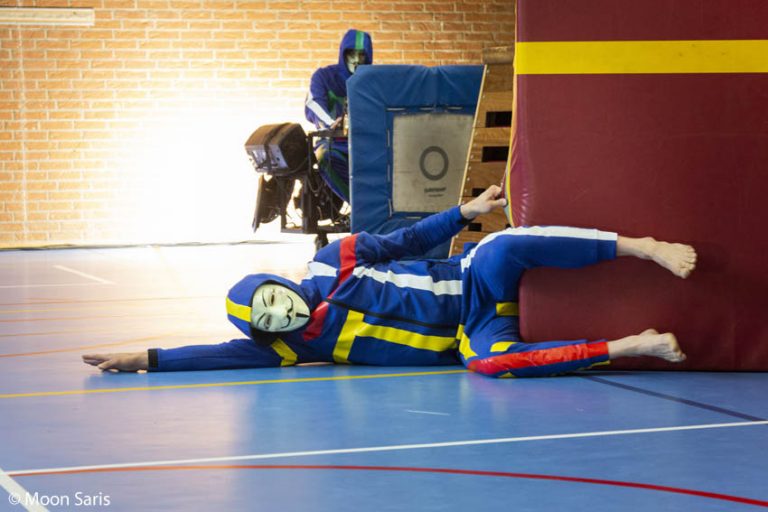 Een persoon gekleed in een blauw en geel trainingsoutfit ligt half achter een trainingshindernis in een sporthal.