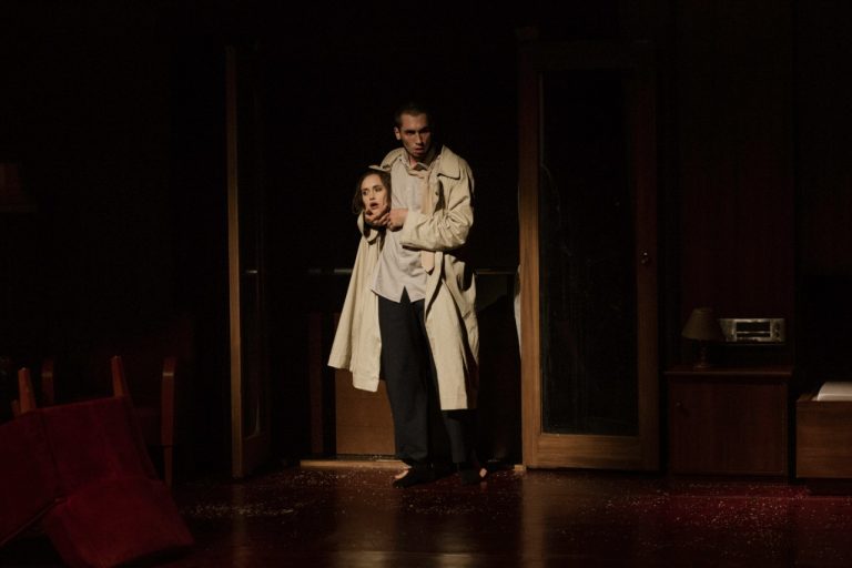 Een personage in een lange jas houdt een hoofd vast, terwijl het in een donkere setting staat.