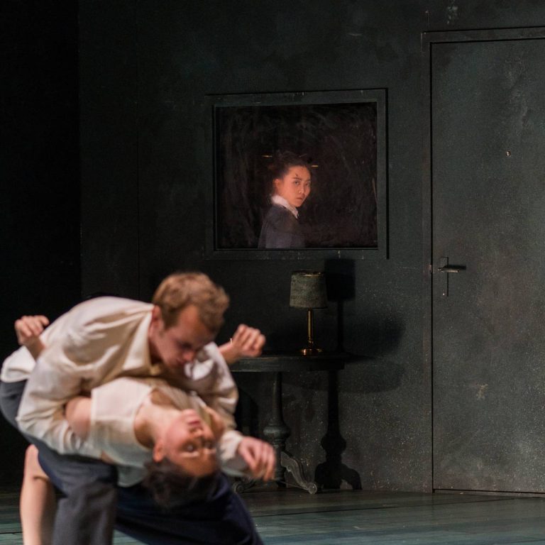 Twee acteurs zijn in een dramatische pose op een toneel, terwijl een derde acteur in een spiegel of kamer daarachter toekijkt.