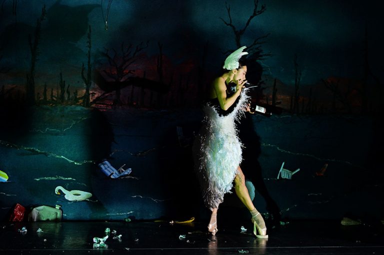 Een danseres in een witte, verenjurk poseert dramatisch tegen een donker, surrealistisch decor.