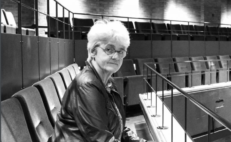 Een oudere vrouw met grijs haar zit op een stoel in een theateromgeving, terwijl ze naar de camera kijkt.
