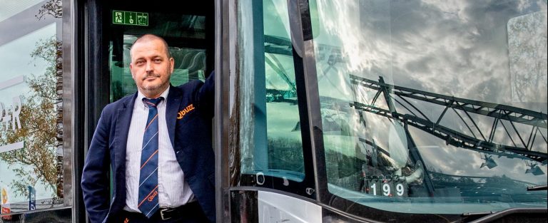 Een man in een uniform staat bij de deur van een bus, met een moderne omgeving op de achtergrond.