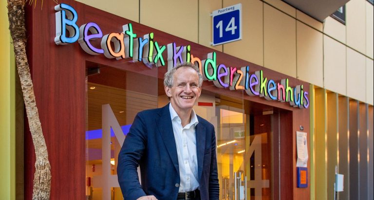 Een man in een blazer staat lachend voor een kleurrijke ingang met de tekst