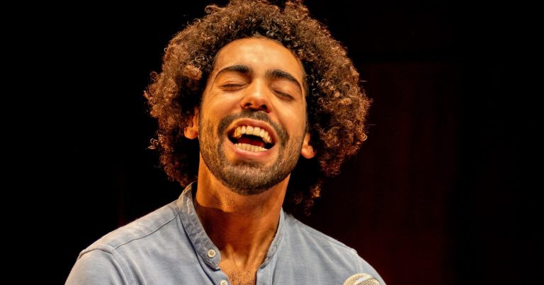 Een man met een volle, krullende haardos zingt enthousiast en glimlacht.