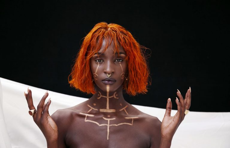 Een persoon met een korte, oranje haarsnit en traditionele bodypaint poseert met beide handen omhoog voor een donkere achtergrond.