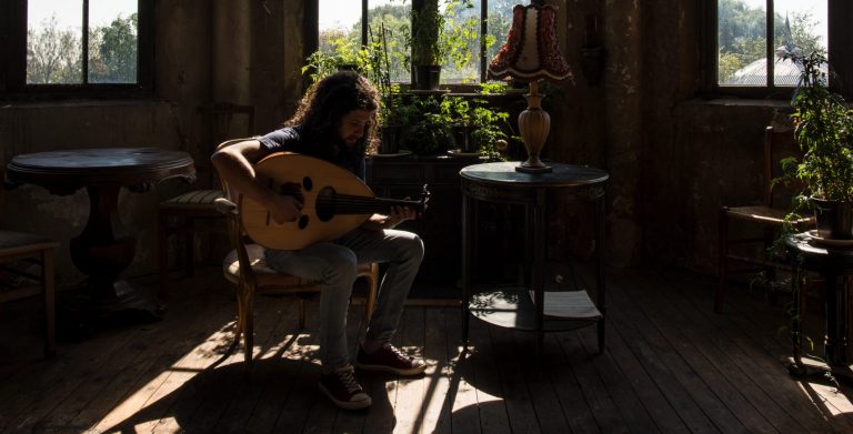 Een persoon zit op een stoel en speelt gitaar in een sfeervolle ruimte met planten en een lamp op een bijzettafel.