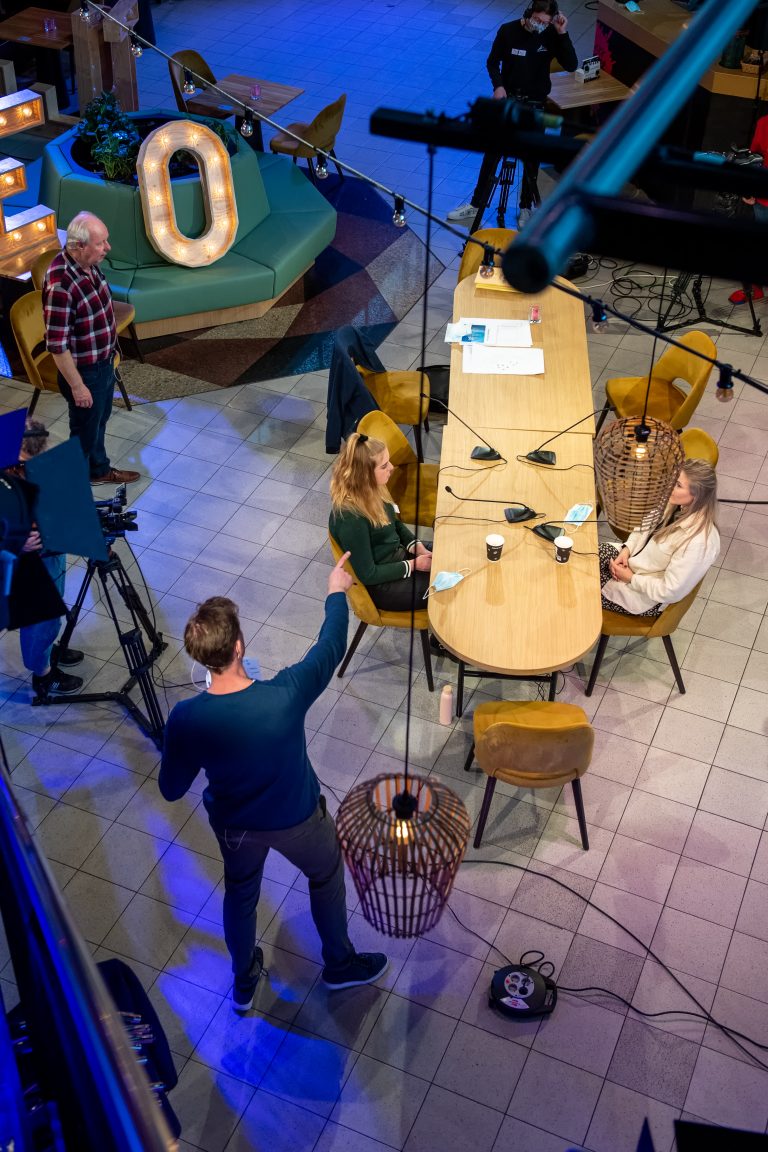 Een overhead opname van een opnamestudio met een lange tafel, waar twee personen zitten en een cameraman bezig is met het filmen.