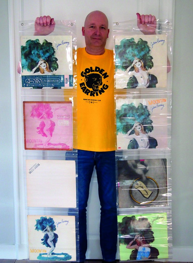 Een persoon staat rechtop voor een muur met gekleurde panelen en draagt een felgeel T‑shirt met een zwart cirkelvormig ontwerp.