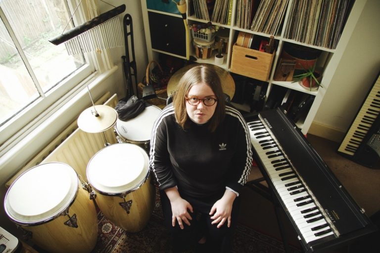 Een persoon zit tussen muziekinstrumenten, waaronder conga's en een piano, in een gezellige kamer met platenwanden.