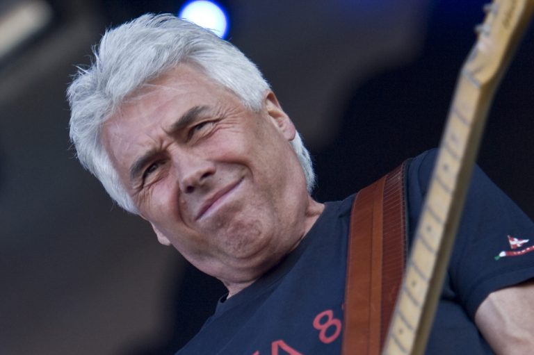 Een man met grijs haar speelt gitaar en heeft een gefocuste uitdrukking op zijn gezicht.