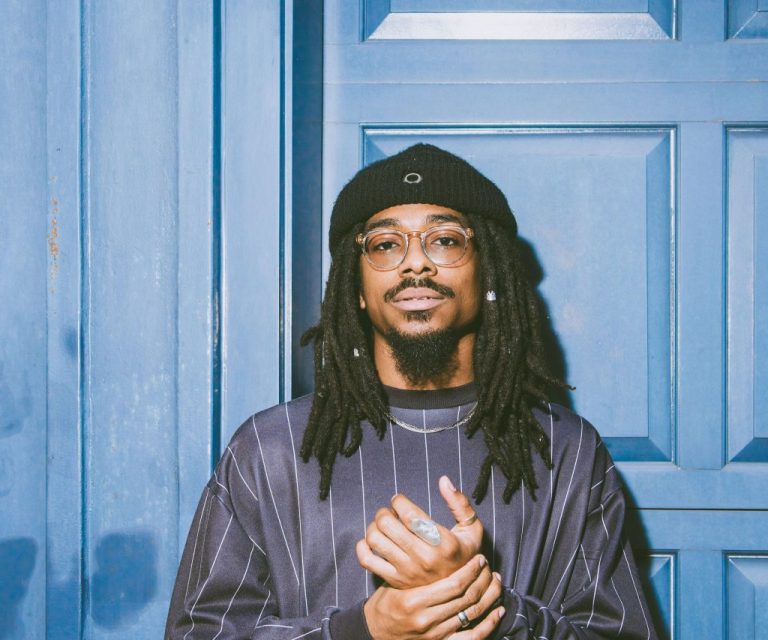 Een man met een zwarte hoed en een gestreepte trui staat voor een blauwe deur.