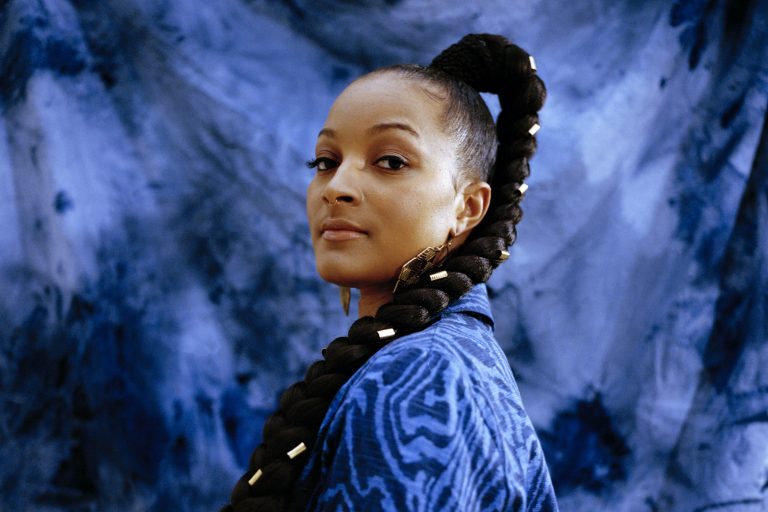 Vrouw met een lange vlecht, gekleed in een blauw patroon, tegen een blauwe achtergrond.