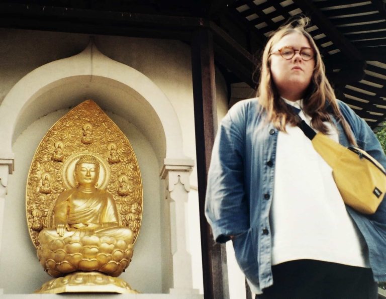 Een persoon staat voor een gouden Boeddha-beeld in een architectonische setting.