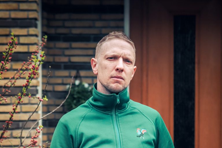 Man in een groene zip-up jacket staat voor een stenen muur met een houten deur op de achtergrond.