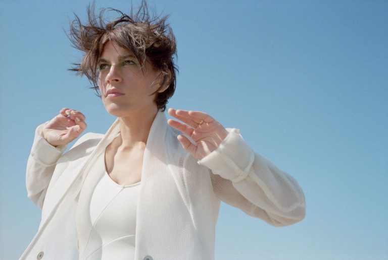 Vrouw met los haar staat in een lichte outfit tegen een blauwe lucht.