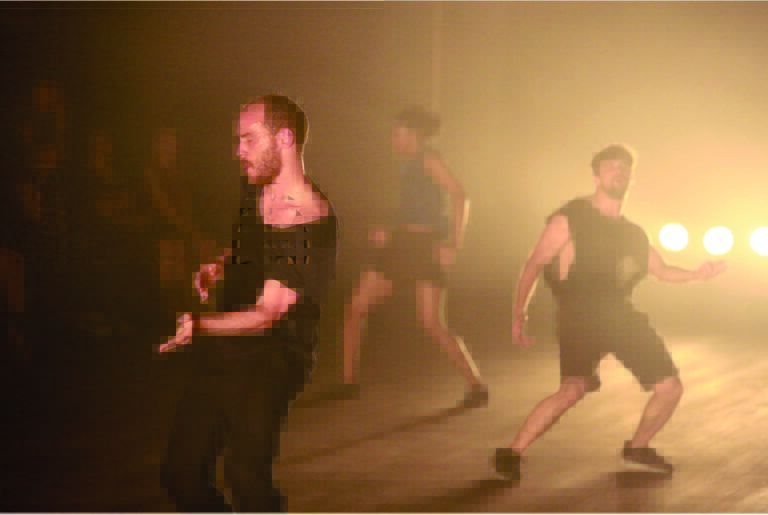 Een groep dansers in zwarte kleding beweegt spectaculair in een mistige, goudkleurige omgeving.