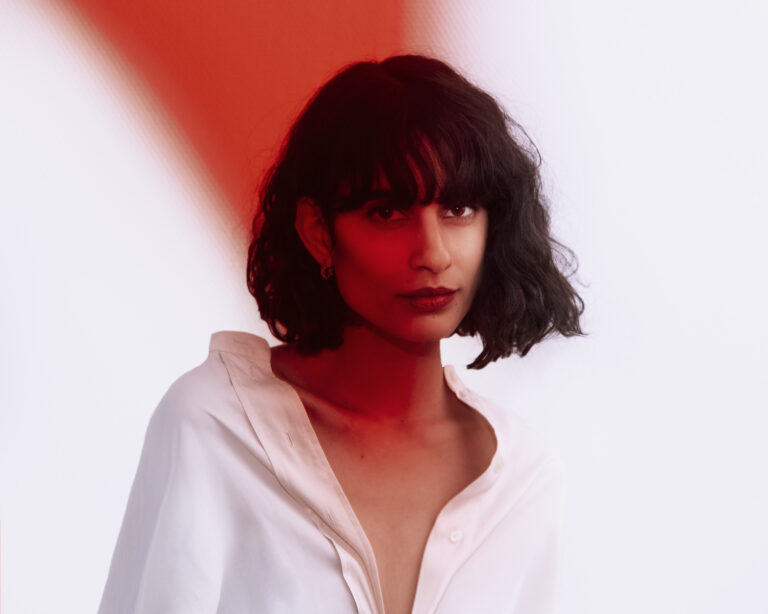 Portret van een vrouw met schouderlang zwart haar, gekleed in een witte blouse, tegen een achtergrond van vage rode en witte kleuren.