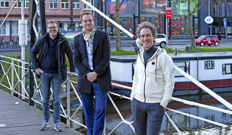 Drie mannen poseren lachend op een brug bij een waterweg.