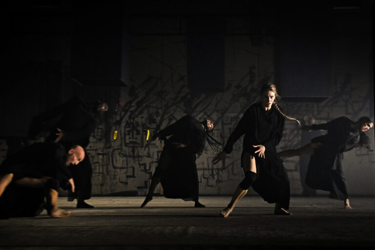 Groep dansers in zwarte kleding die dynamische bewegingen uitvoeren op een donkere achtergrond.