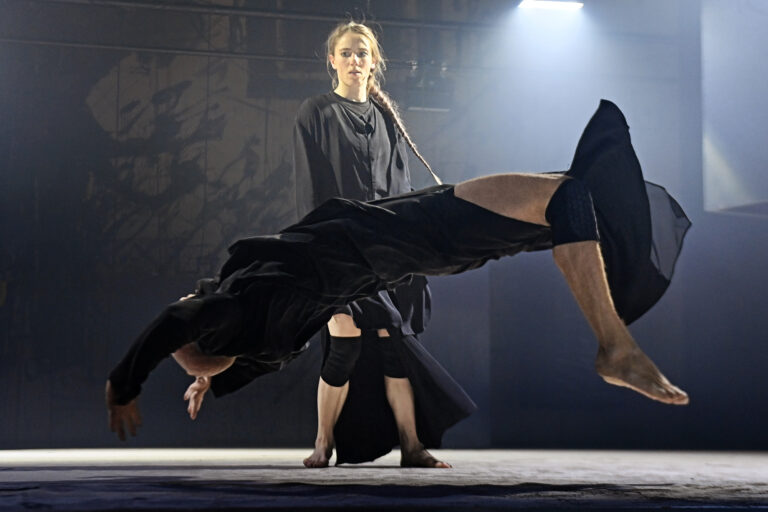 Een danser voert een acrobatische sprong uit terwijl een andere danseres in zwarte kleding toekijkt.