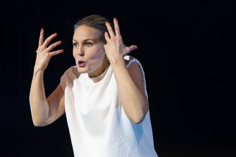 Een vrouw in een witte top maakt een expressieve gebaar terwijl ze op een podium staat.