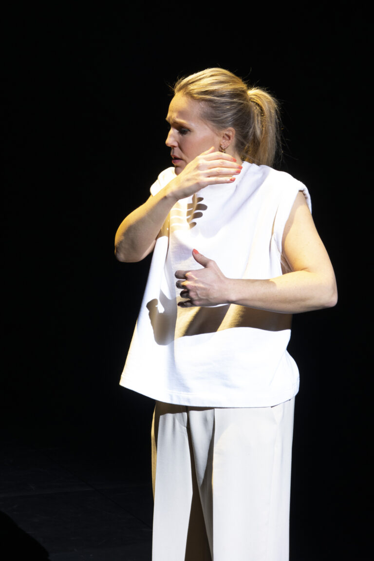 Een vrouw in witte kleding voert een expressieve beweging uit op een podium met een donkere achtergrond.