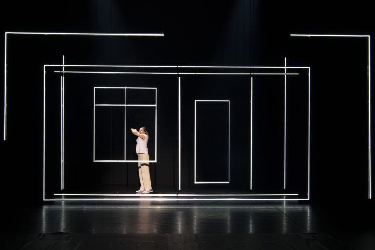 Een persoon staat in een minimalistisch, verlicht decor met grote, geometrische vormen.