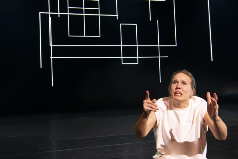 Een danseres in een wit kostuum voert een expressieve beweging uit op een leeg podium met geometrische lijnen op de achtergrond.