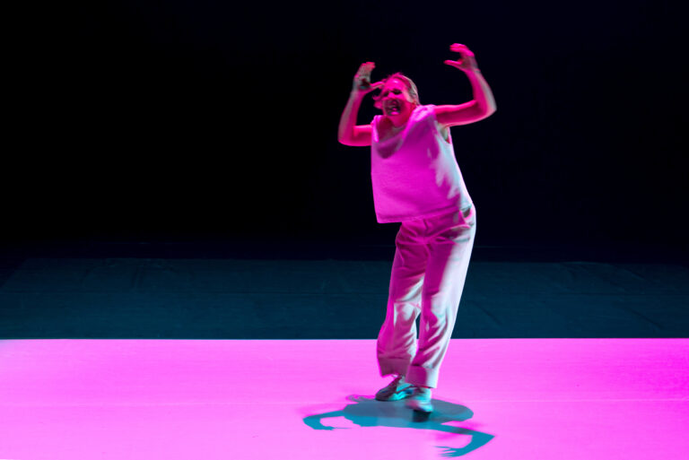 Een danser in witte kleding beweegt expressief op een felroze podium met een donkere achtergrond.