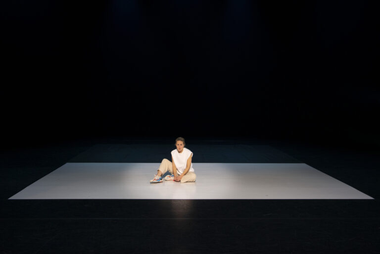 Een danser zit op een lichtgekleurde vloer in een donkere ruimte.