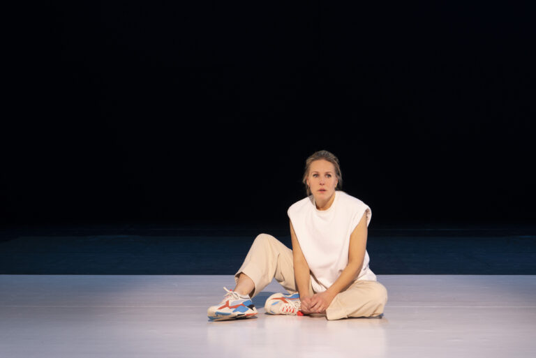 Een danseres zit op de vloer in een minimalistische, donkere ruimte.