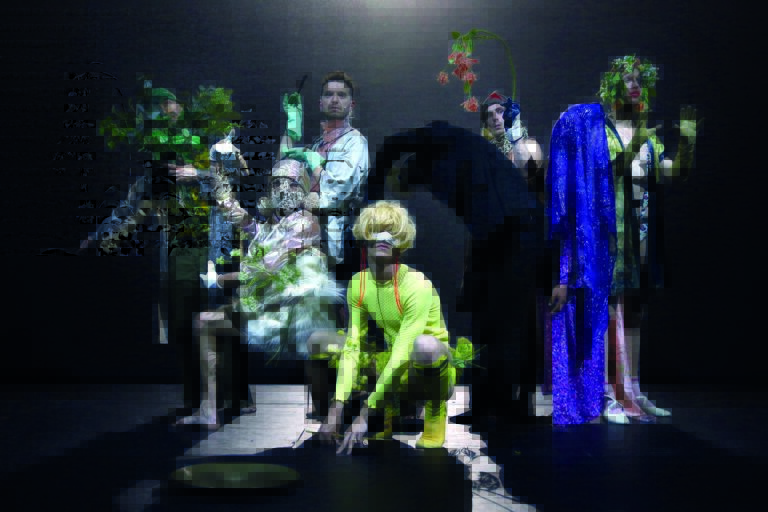 Een artistieke groep van mensen in kleurrijke kostuums en scènes, omgeven door planten en decor.