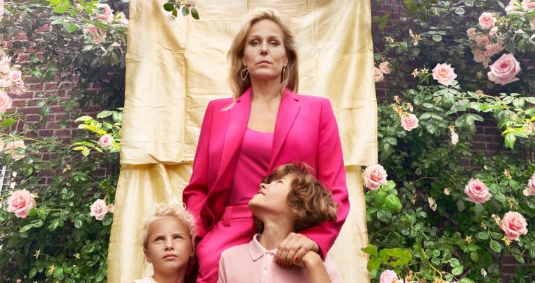 Een vrouw in een felroze pak poseert voor een gouden achtergrond, terwijl twee kinderen naar haar kijken.